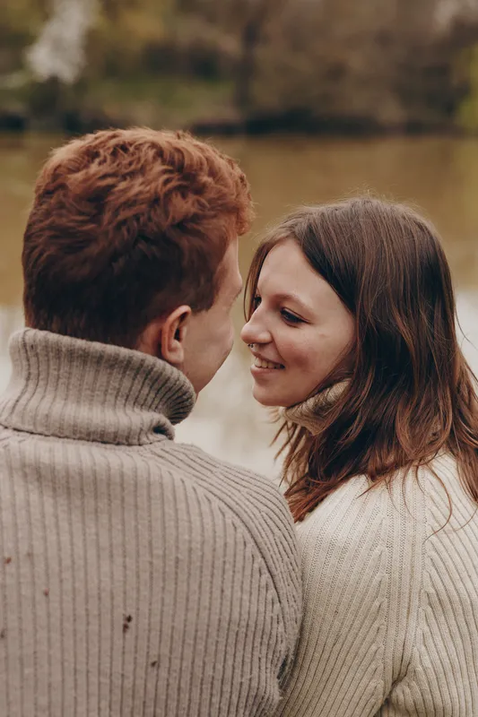 A young couple wearing cozy knit sweaters shares a tender moment outdoors near a calm river in autumn, expressing warmth and connection in a natural lifestyle setting.