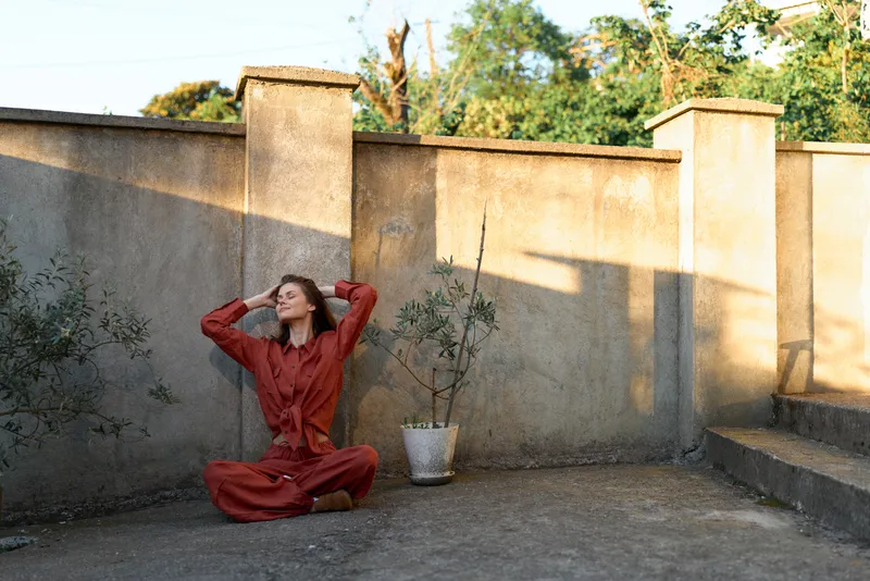 Woman sits on a sunlit terrace wall in a red dress, hands in hair, calm relaxed pose, urban lifestyle portrait with clear natural light and warm mood for lifestyle