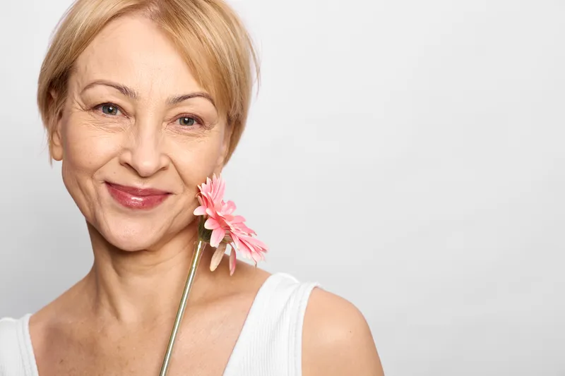 Woman portrait, older blonde smile, pink flower on stick, soft studio light on face, beauty and skincare concept, calm confident wellbeing, with natural femininity today
