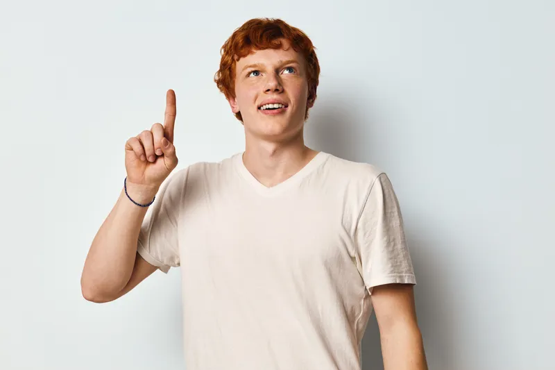 Young man with red hair and white t shirt pointing up with index finger, looking away and smiling on plain light background in studio. Casual style, positive expression.