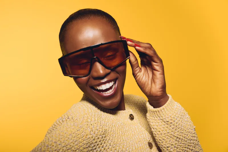 Smiling African American woman with dark skin wearing large sunglasses and a beige knitted sweater on yellow background. Concept of fashion, happiness, style, and confidence.