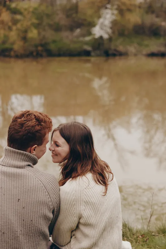 Romantic couple sits close by a calm lake in autumn, exchanging smiles and a tender glance, wearing cozy sweaters, natural backdrop, warm light, candid lifestyle moment by water, and soft wind.