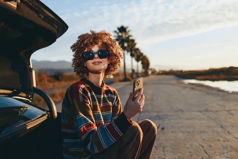 Woman lifestyle outdoors, curly hair and sunglasses, sitting in an open car doorway on a road, holding a smartphone, warm sunset light, casual travel mood, soft cinematic blur
