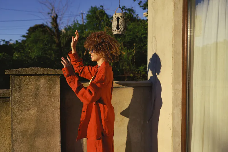 Casual woman lifestyle shot outdoors, a curly haired woman in a bold orange outfit raises arms on a sunny balcony, expressing playfulness and freedom against a concrete wall in warm afternoon light