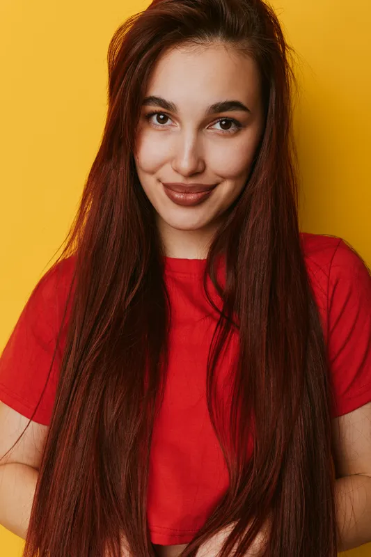 Young woman with long straight hair and red shirt smiling confidently against yellow background. Portrait of happy female with natural makeup and relaxed pose in studio lighting.