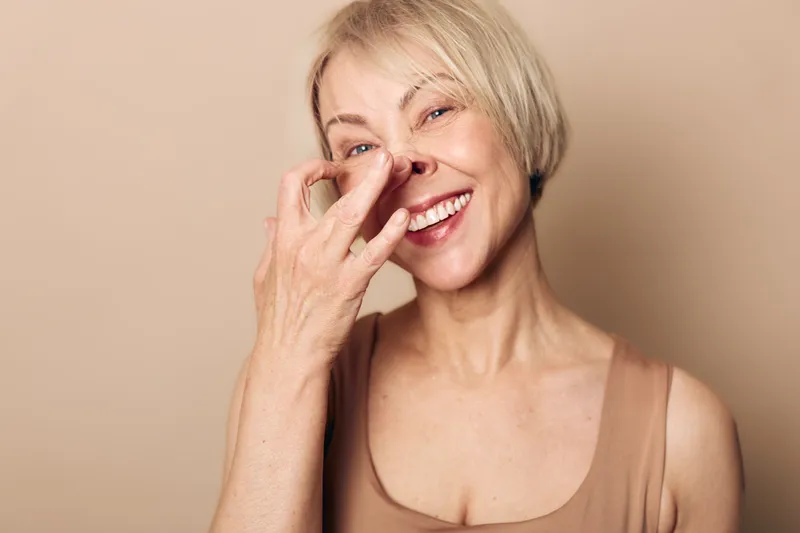 Happy mature woman with short hair smiling and holding nose in playful gesture. Neutral background and casual outfit create relaxed and humorous portrait concept.