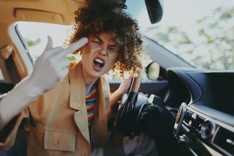 Woman with curly hair inside a car, shouting with a fierce expression, making a bold gesture, capturing a rebellious lifestyle moment.