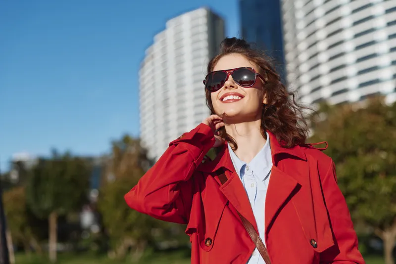 A stylish young woman in a red trench coat smiles as she adjusts her sunglasses in a sunny urban park, with modern high-rise buildings behind her.