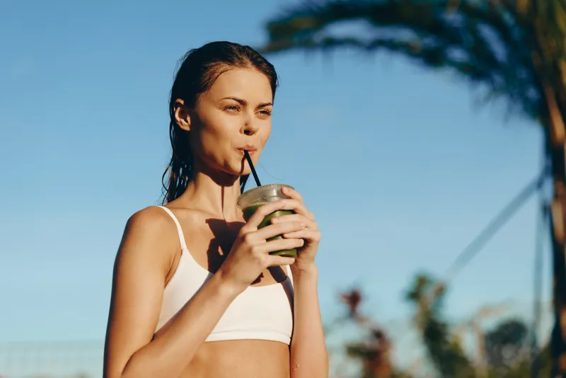 Young woman in a white crop top outdoors drinks a smoothie through a straw, enjoying a sunny day, palm trees behind her, casual fitness vibe, bright summer lifestyle.