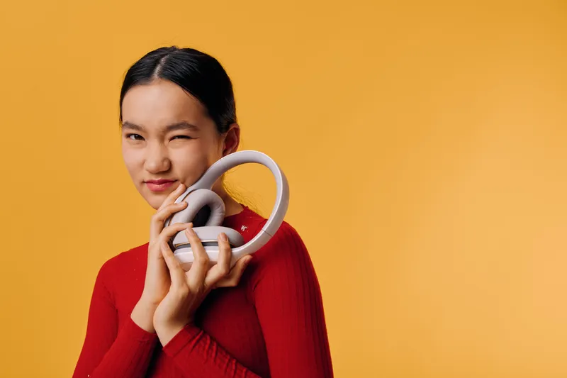 Young woman in red sweater holding white headphones close to her face, smiling with one eye closed against orange background. Relaxed, casual style with cozy mood and tech accessory.