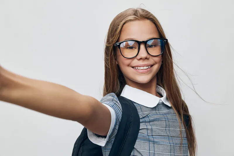 Girl smiling with long hair wearing glasses and school uniform holding backpack taking selfie on gray background. Child happy with education and style