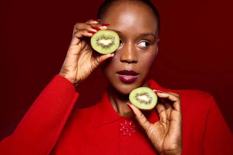Dark skinned African woman in red sweater holds two kiwi halves near her face against red background. Concept of healthy food, beauty, and natural skincare with fresh fruit and vibrant colors.