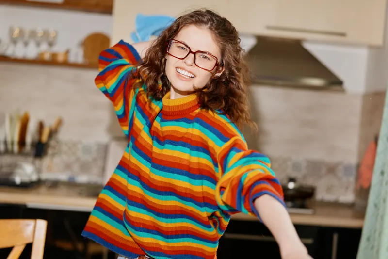 Cheerful woman indoors, wearing a colorful striped sweater, smiling with glasses, casual home moment, warm lighting, cozy kitchen backdrop, relaxed pose with natural textures