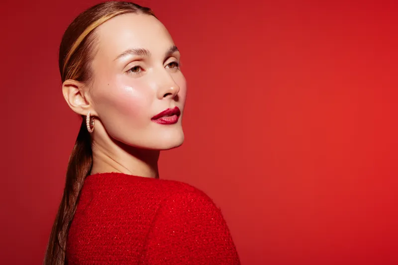 Portrait of a confident woman with shiny skin and red lips wearing a red sweater against a red background. Concept of beauty, fashion, skincare, and elegance in studio lighting.