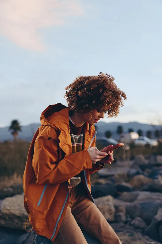 Woman with curly hair sitting outdoors on rocky ground, orange jacket and backpack, checking a smartphone during travel at sunset, blurred nature and mountains, calm lifestyle mood