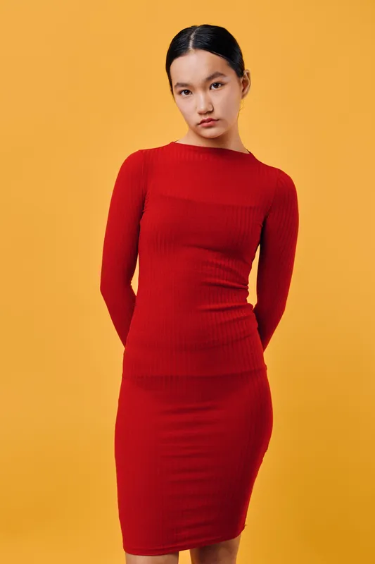 Young woman wearing red dress standing against orange background, looking at camera with neutral expression. Studio portrait with simple colorful backdrop and casual posture for fashion or style
