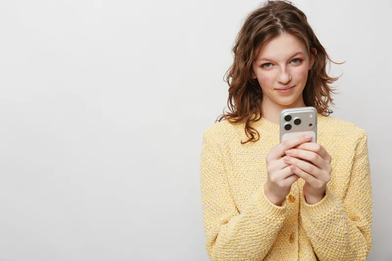 Young woman in yellow sweater holds smartphone with both hands and looks at camera with slight smile. Concept of communication, technology, social media, and casual lifestyle.