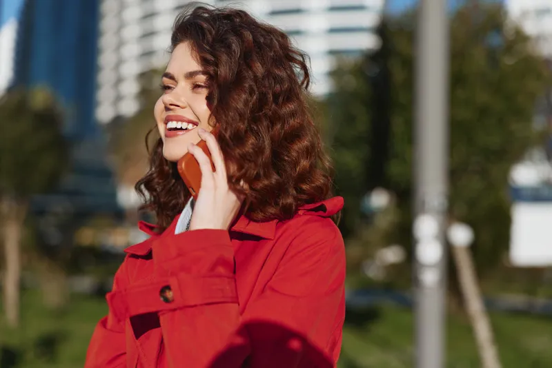 Young woman with curly hair in a red coat chats on a smartphone in a sunny urban park, smiling and engaged during a casual outdoor call, enjoying the moment.