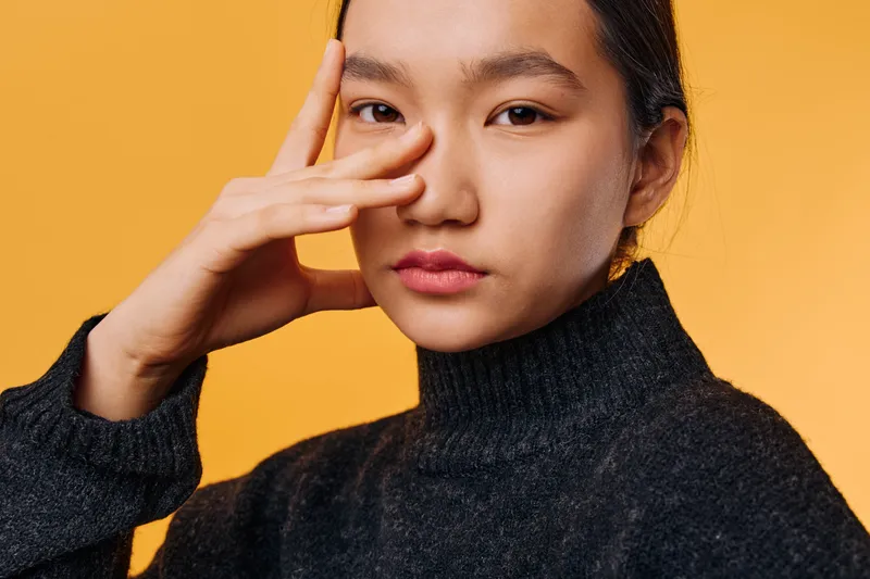 Young woman in black sweater posing with thoughtful expression and hand near face on orange background. Closeup portrait of asian female with natural makeup and neat hairstyle indoors.