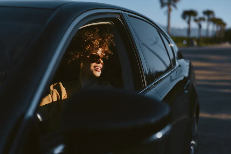 A smiling woman with curly hair sits in a car window enjoying a sunset drive, captured in a retro film color mood with warm tones and nostalgic atmosphere.