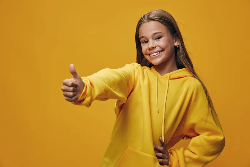Girl in yellow hoodie smiling and showing thumbs up gesture with one hand on waist against bright yellow background. Positive expression and casual style for youth and happiness concept.