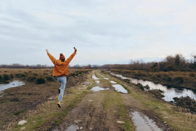Cheerful woman lifestyle outdoors, running on a muddy path beside a calm river in early spring, arms lifted, dynamic pose, overcast daylight, natural countryside colors, grass and trees