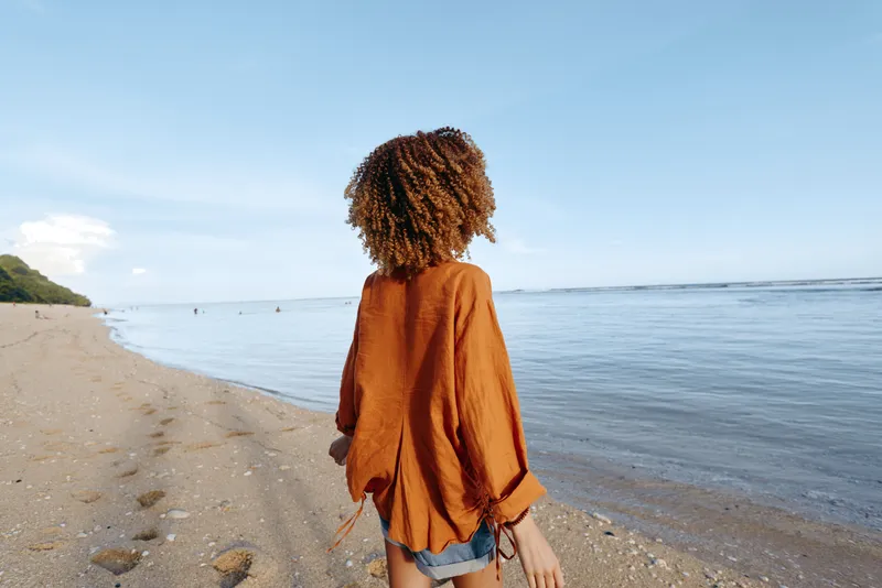Beach scene, woman standing on sandy shore, backview to camera, calm ocean horizon, quiet waves, person outdoors under blue sky, relaxed posture, summer travel mood, natural colors