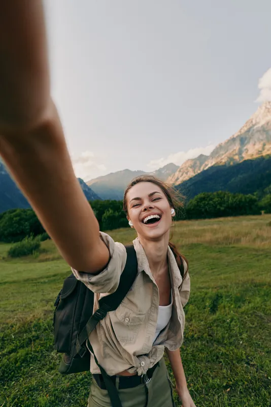 Playful young woman outdoors reaches toward the camera with a big smile as she poses on a grassy meadow, a backpack resting on her shoulders, mountains looming in the distance