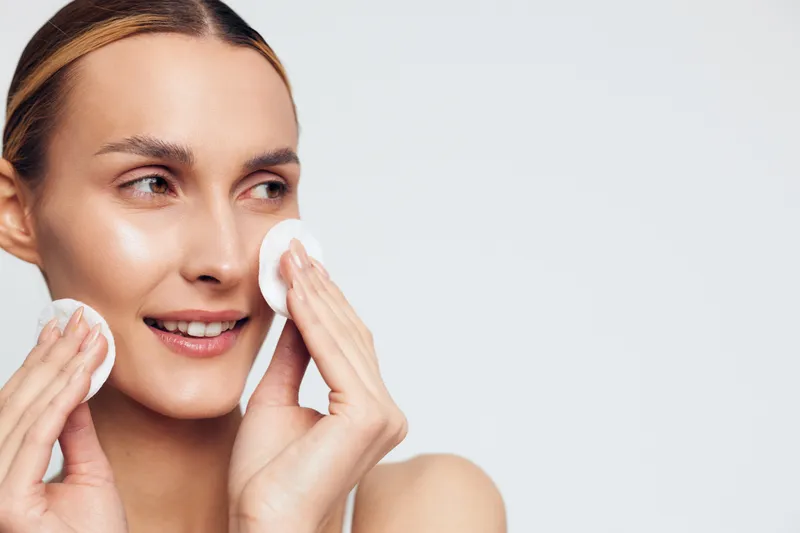A young woman with clear skin uses cotton pads to gently cleanse her face against a plain background. The image represents skincare routine and natural beauty care concepts.