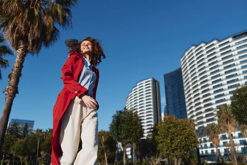 Cheerful woman outdoors wearing a red coat, standing in a sunlit urban park with palm trees and tall buildings, conveying confident style and breezy motion. Ambient energy.