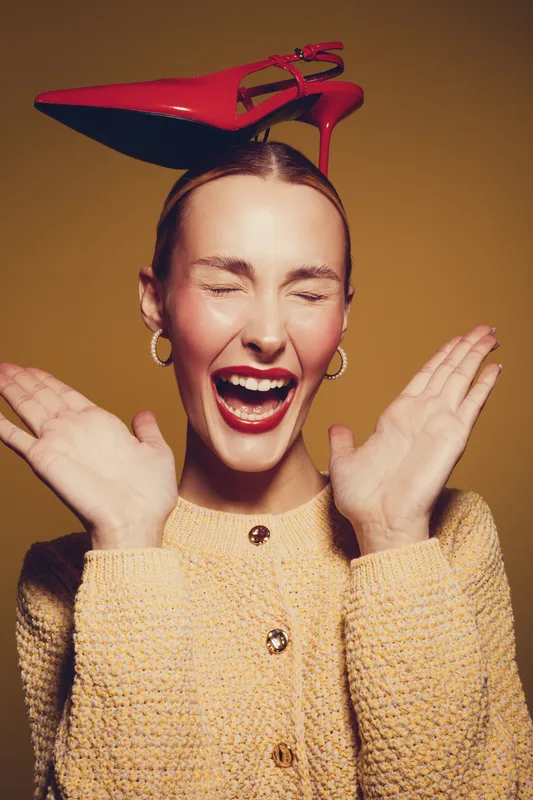 Happy woman with closed eyes and red lipstick laughs joyfully wearing yellow sweater and red high heel shoe on head. Concept of fun, fashion, and playful mood in studio lighting.