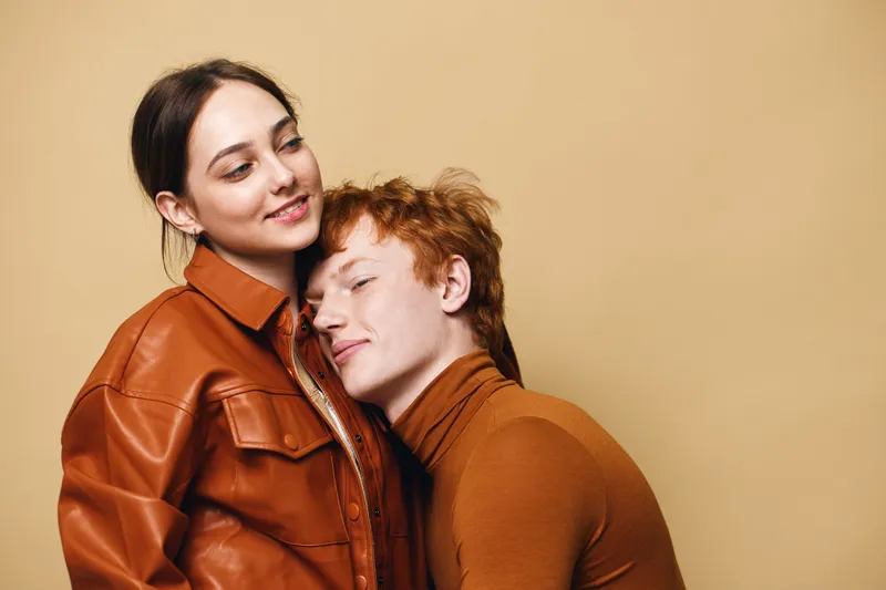 Young couple with affectionate expression, woman in brown leather jacket and man with red hair in brown turtleneck hugging gently on beige background indoor studio.