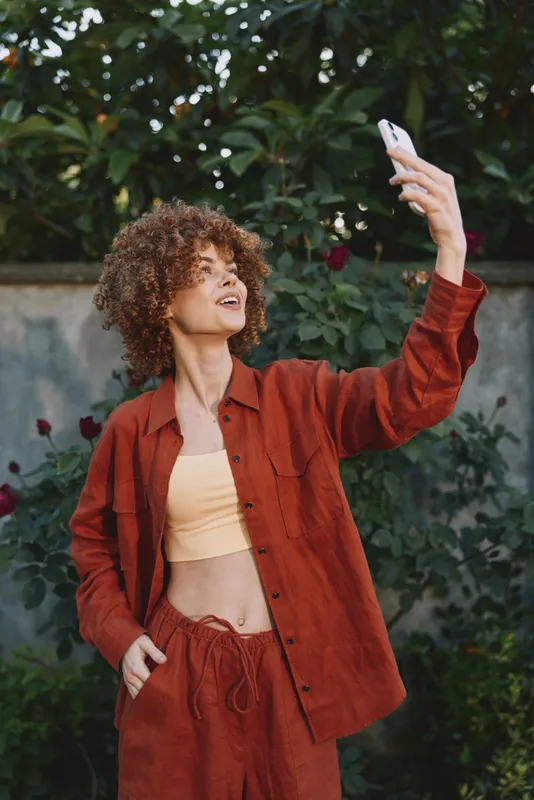 A stylish woman takes a carefree selfie in a sunlit garden, wearing a coordinated rust outfit. Casual mood, fashion forward vibe, and confident pose for lifestyle and self expression
