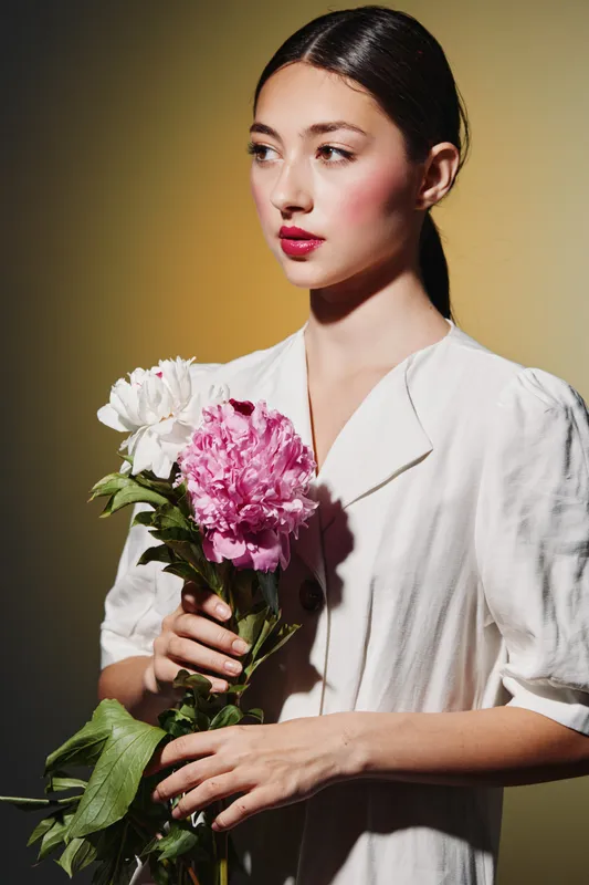 Young woman in stylish white blouse holding pink and white flowers looks aside thoughtfully. Studio shot with strong shadows and warm background. Concept of beauty, fashion, and mood.