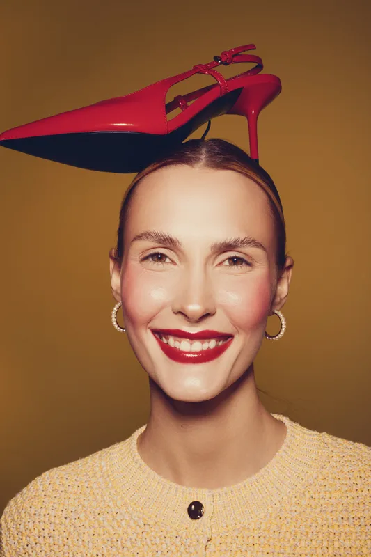 Smiling woman with red high heel shoe balanced on her head wears yellow sweater and hoop earrings. Studio portrait on brown background. Concept of fashion, style, and playful mood.