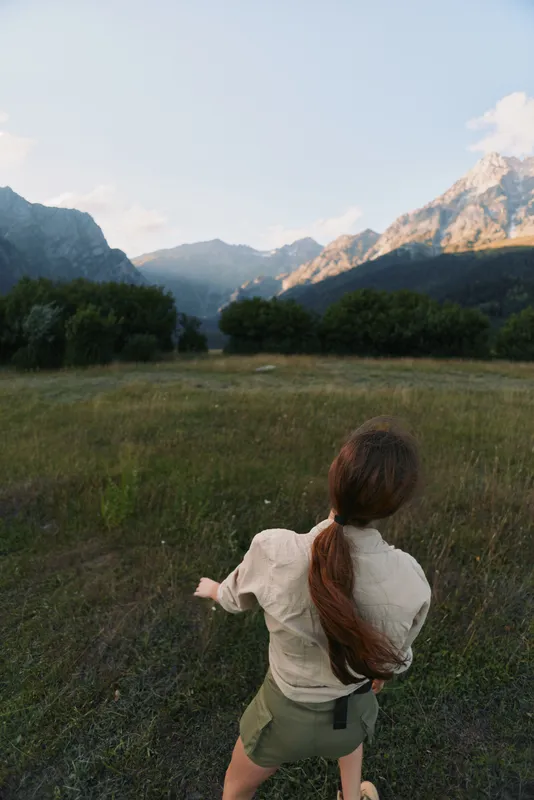 Young woman with a ponytail walks through a sunlit meadow toward distant mountains under a clear sky, conveying exploration, freedom, and a sense of open possibility.