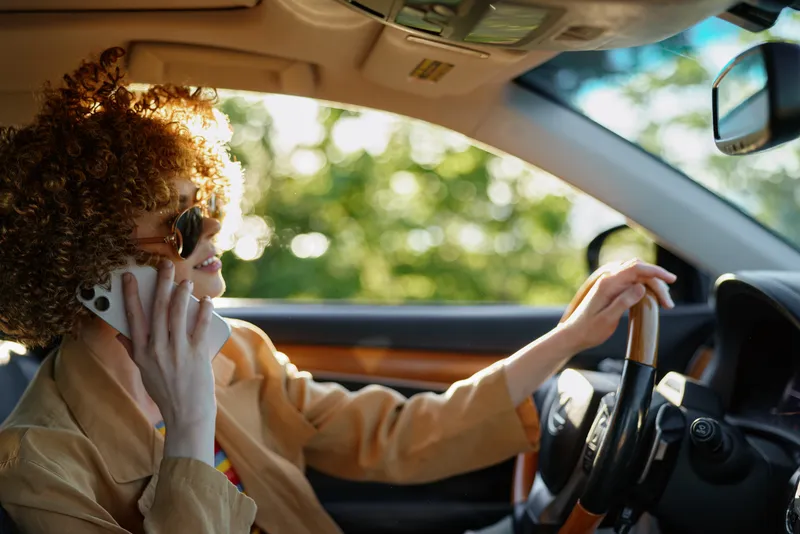 A woman driver sits in a warm car interior, talking on a mobile phone while steering. Natural light highlights curly hair. Concept for urban commute and retro lifestyle.