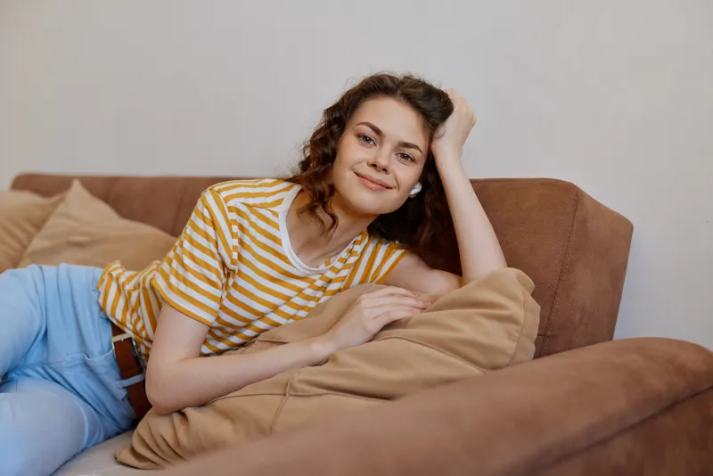 A relaxed woman lies on a brown couch at home, smiling with her head resting on a pillow. A casual lifestyle scene highlighting comfort, self care, and everyday moments in a bright living space.