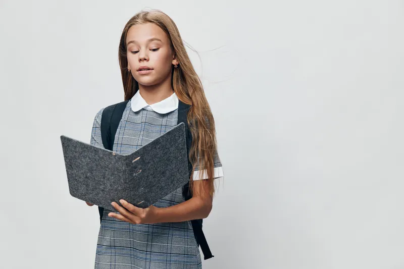 Teenage girl reading from folder with focus, standing isolated on white background. Student with backpack in casual outfit practicing presentation or speech for school assignment.