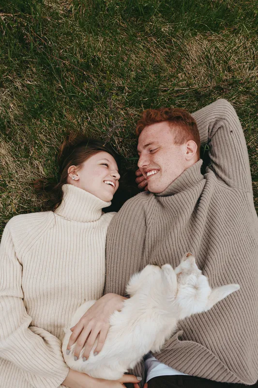 Cheerful couple outdoors, lying on grass in cozy sweaters, warm sunlight and gentle breeze, relaxed smiles, intimate lifestyle moment, natural colors, soft textures, candid connection, peaceful mood,