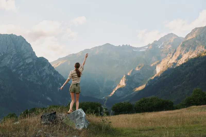 Woman outdoors on a rock in a meadow, arms raised in a carefree pose, majestic mountains in the background, sunlit scene, natural colors, lifestyle moment