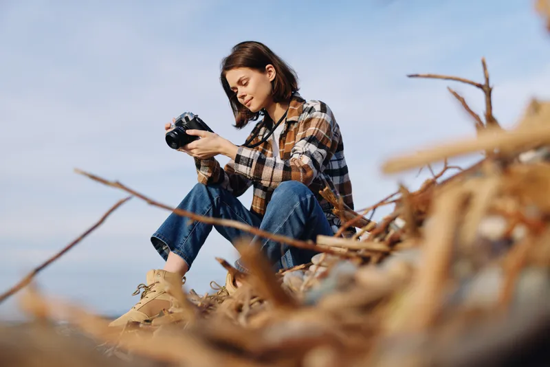 Woman photographer sits on a rocky shore, plaid shirt and jeans, focused on her camera as she captures the sea, sky and rugged landscape with calm outdoor energy.