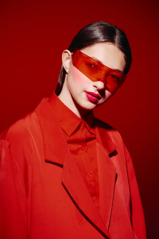 Stylish young woman in oversized red coat and matching transparent red sunglasses poses against vivid red background. Beauty fashion concept with creative colorful style.