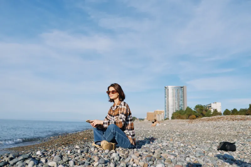 Beach scene with a casually dressed woman sitting on pebbled shore by the sea, holding a phone, clear sky and distant city skyline creating a relaxed travel mood.
