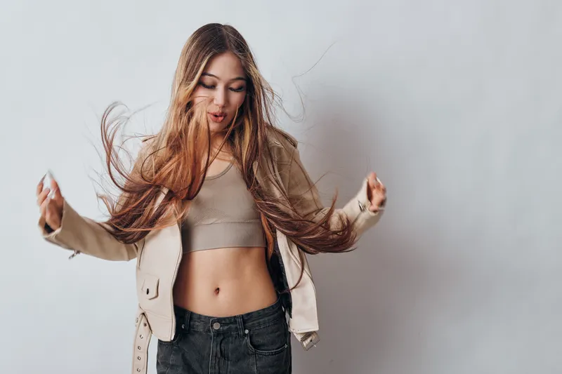 Young woman with long hair in casual clothing dancing and enjoying herself in front of gray wall. Lifestyle, youth, motion, happiness, and freedom in fashion portrait.