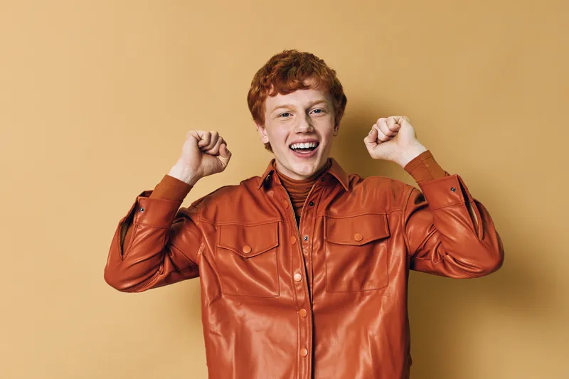 Young man with red hair wearing orange leather jacket smiling and celebrating with raised fists on beige background. Portrait, happiness, emotion and casual clothing style.