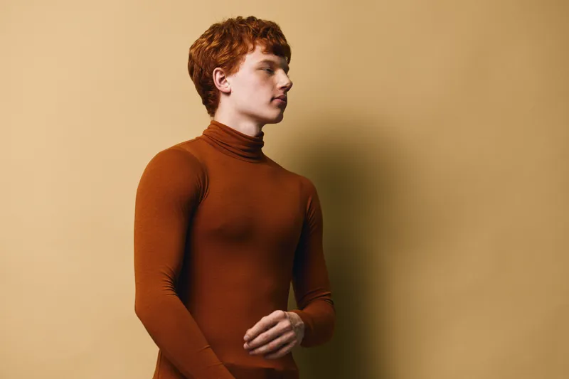 Young man in brown turtleneck standing against beige background looking sideways with serious expression and casual hairstyle in studio portrait for fashion concept.