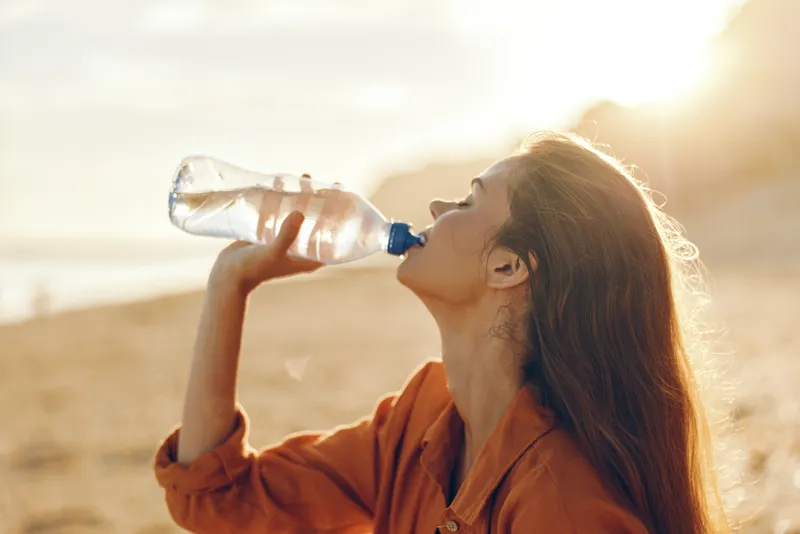 A woman drinks water from a bottle on a sunlit beach, hair blown by the wind, warm light in the sky. Concept for summer hydration and outdoor wellness.