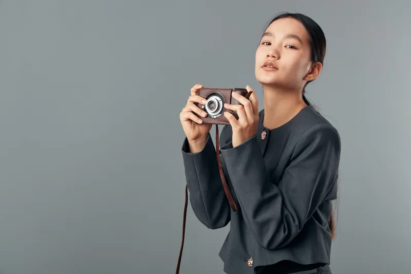 Young woman holding vintage camera with both hands portrait on neutral background. Female person preparing to take photo with retro photographic equipment, casual clothes and natural look.