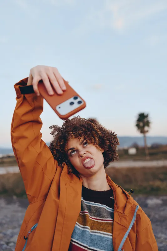 Woman lifestyle outdoors, smiling with curly hair, holding a smartphone for a selfie, wearing an orange jacket, in soft bright sunlight, relaxed pose, open sky background